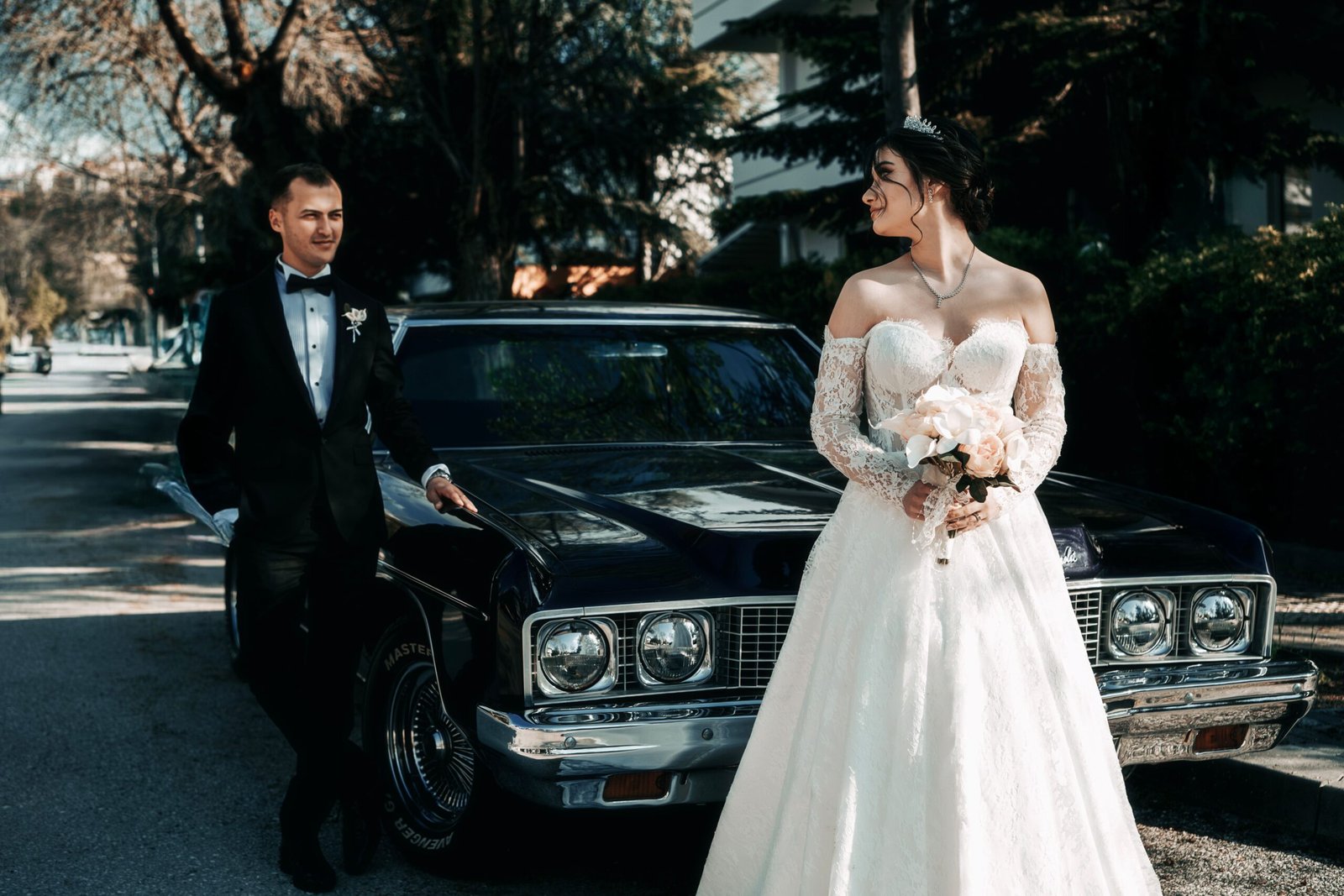 Bride and groom in wedding attire posing with vintage car on a sunny day.