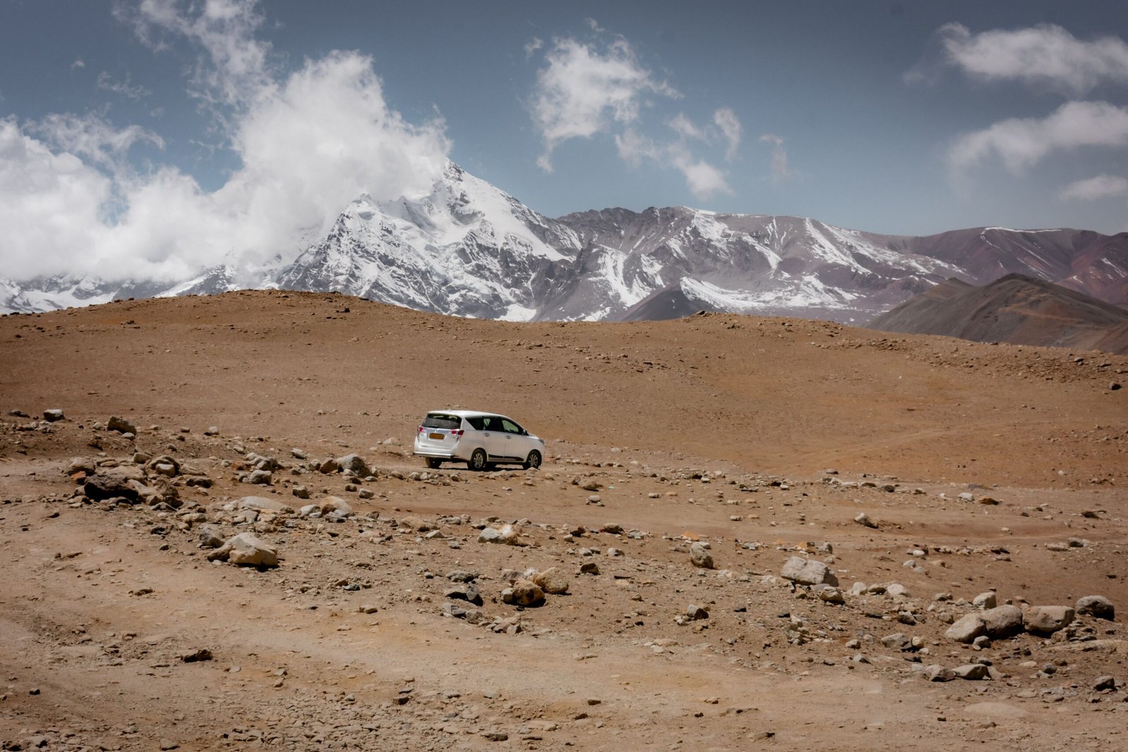 A car navigates the arid desert terrain with snowcapped Himalayan mountains in the background.